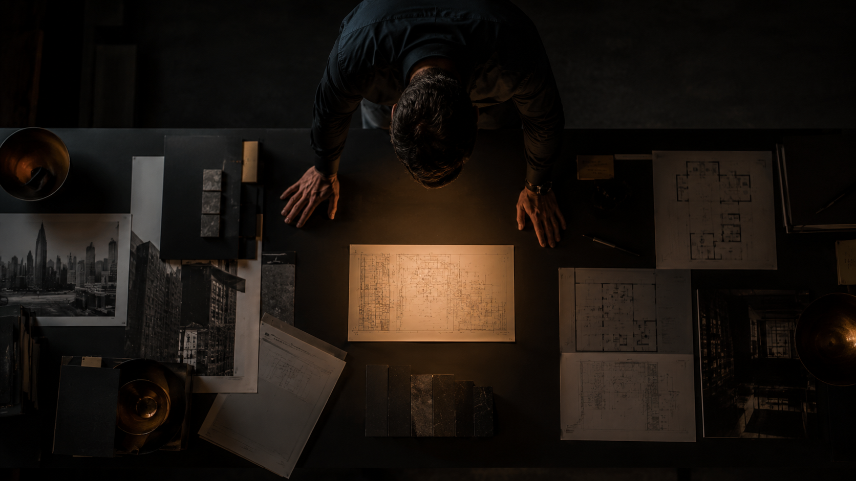 Architect leaning over a desk, examining a central blueprint surrounded by photos, plans, and material swatches.
